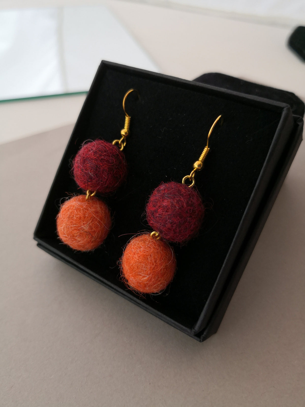 Pair of 14 karat gold earrings with red and orange hand-felted wool balls in a black box on a light surface.